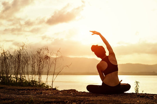 Woman stretching in a seated yoga pose at sunrise beside a calm lake, highlighting the peaceful start of a morning wellness routine.